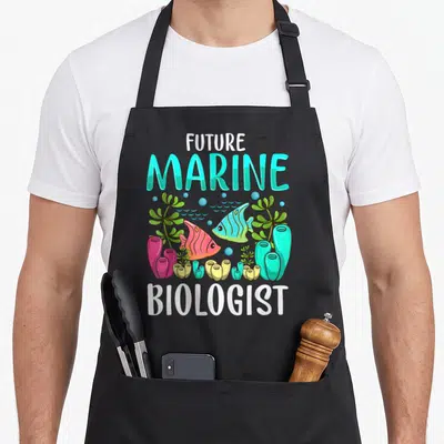 Ocean marine biologist apron - FUTURE MARINE BIOLOGIST text with cartoon fish, coral, and seaweed in cyan and pink — shown on a black cotton canvas apron against a white background