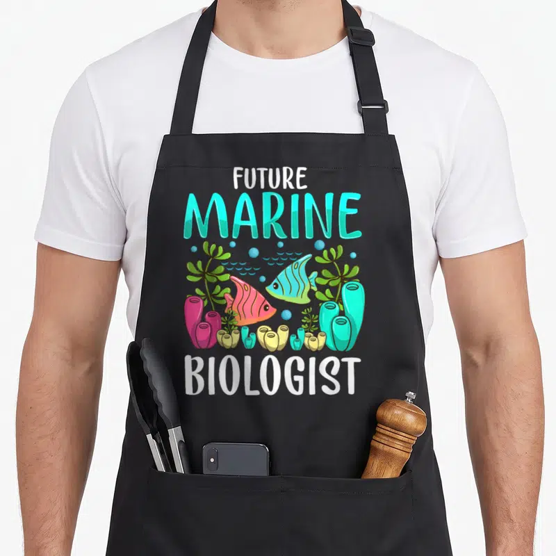 Ocean marine biologist apron - FUTURE MARINE BIOLOGIST text with cartoon fish, coral, and seaweed in cyan and pink — shown on a black cotton canvas apron against a white background
