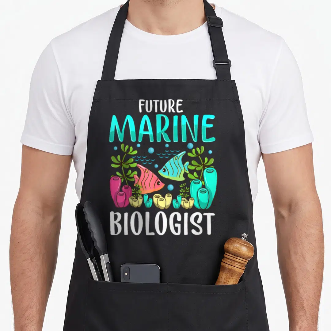 Ocean marine biologist apron - FUTURE MARINE BIOLOGIST text with cartoon fish, coral, and seaweed in cyan and pink — shown on a black cotton canvas apron against a white background