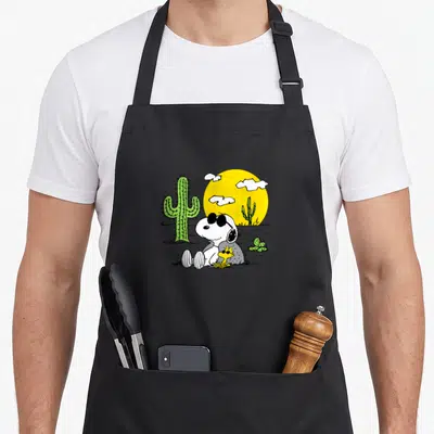 Desert summer aprons - Snoopy and Woodstock sitting near a green cactus under a yellow sun with white clouds — shown on a black cotton canvas apron against a white background