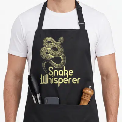 Snake animal whisperer apron - Pale yellow coiled snake illustration above stacked text reading Snake Whisperer — shown on a black cotton canvas apron against a white background