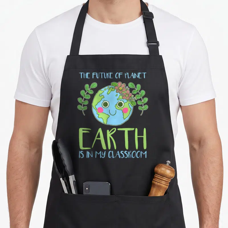 Earth classroom teacher apron - Smiling cartoon Earth with pink flowers and green leaves, flanked by leaf branches — shown on a black cotton canvas apron against a white background