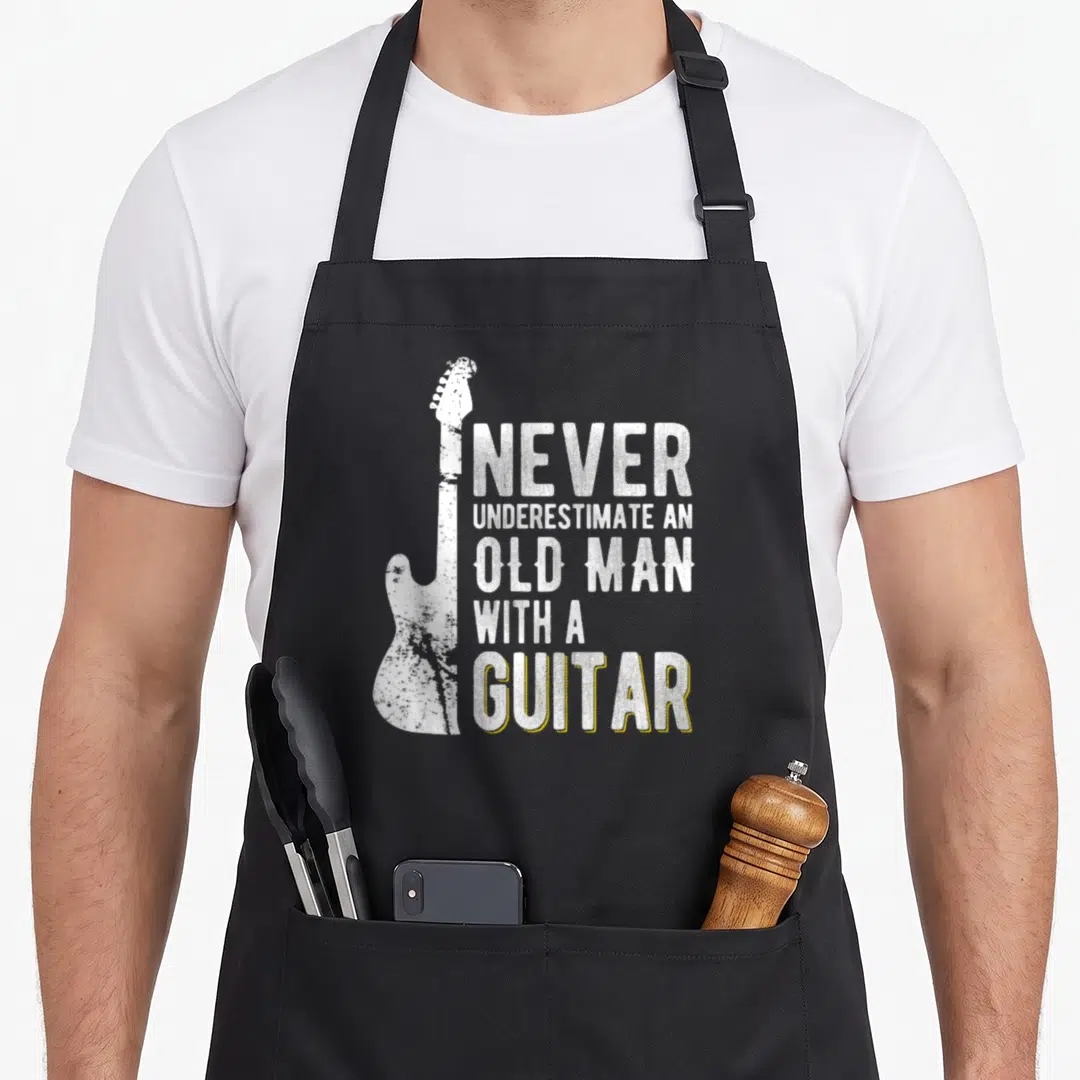 Underestimate guitarist apron - Vertical white distressed electric guitar next to stacked text with yellow drop shadow — shown on a black cotton canvas apron against a white background