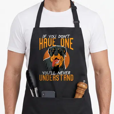 Rottweiler dog breed aprons - Realistic dog head in orange circle with stacked text IF YOU DON'T HAVE ONE, YOU'LL NEVER — shown on a black cotton canvas apron against a white background