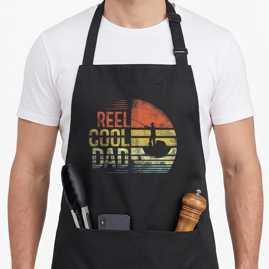 Daddy fisherman apron - Retro REEL COOL DAD text with fisherman silhouette and striped sun in orange and yellow — shown on a black cotton canvas apron against a white background