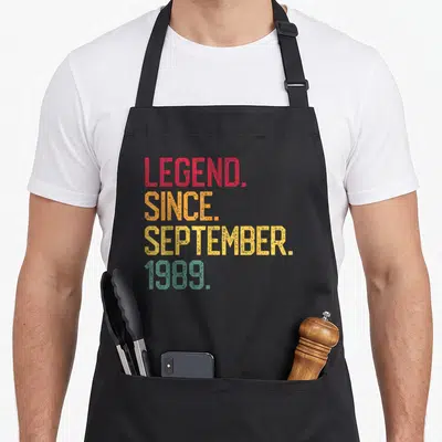 September 36th birthday apron - Stacked red orange yellow teal text reading LEGEND SINCE SEPTEMBER 1989 on black fabric — shown on a black cotton canvas apron against a white background