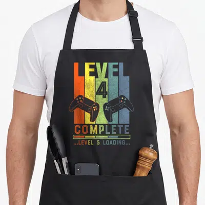 Level complete wedding apron - Retro game controllers on orange yellow green blue purple stripes with leveling text — shown on a black cotton canvas apron against a white background