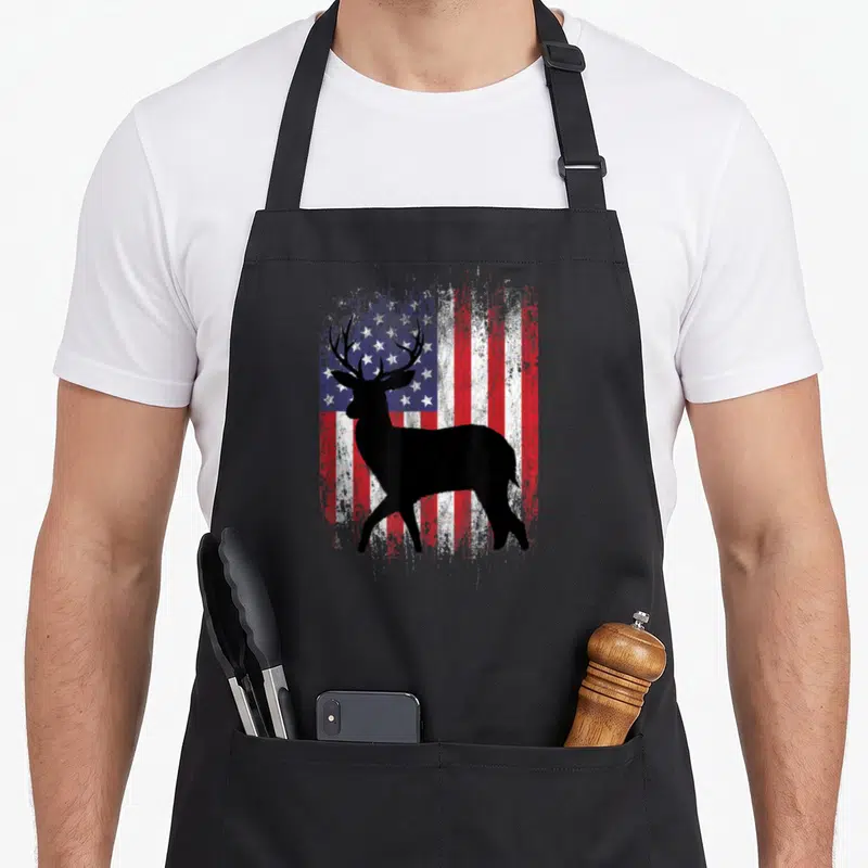 Deer hunting grill hunter apron - Black deer silhouette standing before a distressed red white and blue American flag — shown on a black cotton canvas apron against a white background