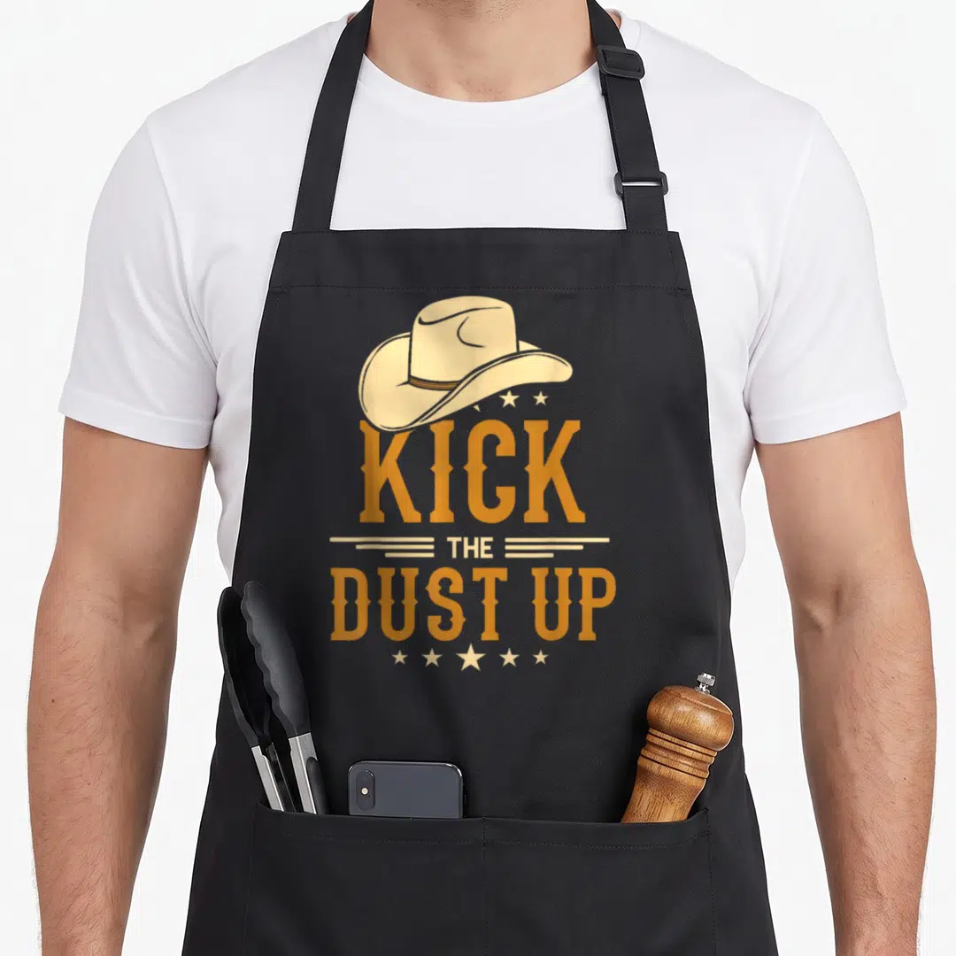 Kick cowboy apron - Beige cowboy hat above orange KICK THE DUST UP text flanked by horizontal lines and two rows — shown on a black cotton canvas apron against a white background