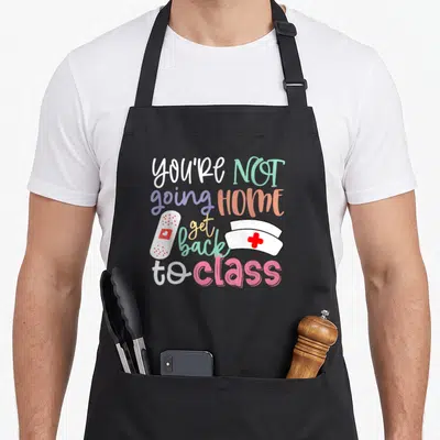 Duty nurse apron - Colorful You're Not Going Home Get Back to Class text with cartoon nurse hat and bandage — shown on a black cotton canvas apron against a white background