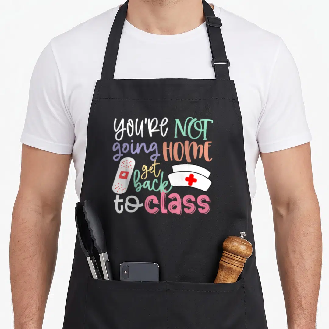 Duty nurse apron - Colorful You're Not Going Home Get Back to Class text with cartoon nurse hat and bandage — shown on a black cotton canvas apron against a white background