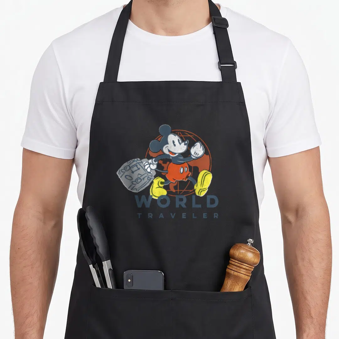 Traveler friends apron - Mickey Mouse carrying suitcase in front of globe with WORLD TRAVELER text — shown on a black cotton canvas apron against a white background
