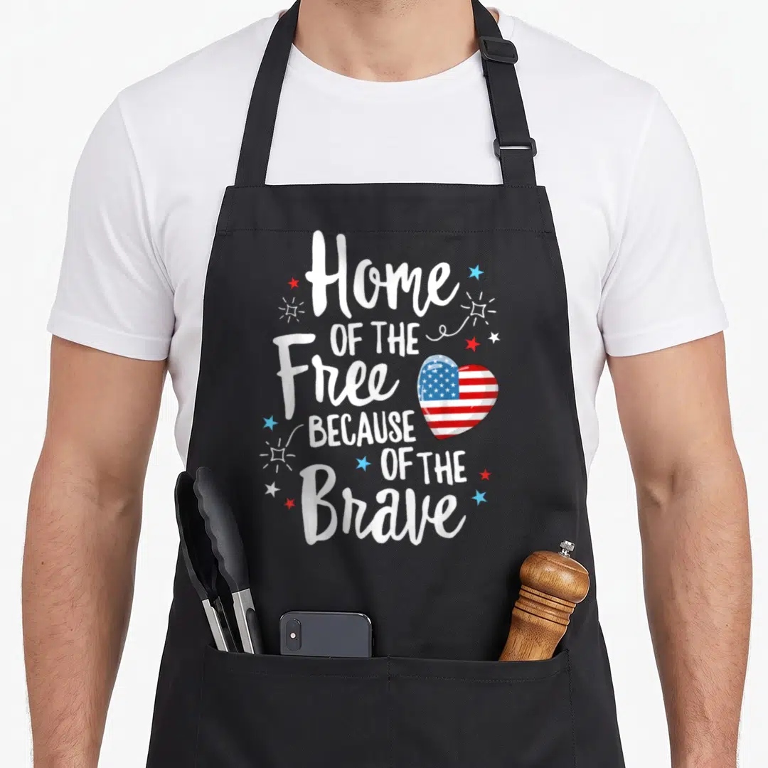 Wife 4th of july apron - Stacked white text Home OF THE Free Because OF THE Brave next to a heart shaped US flag — shown on a black cotton canvas apron against a white background
