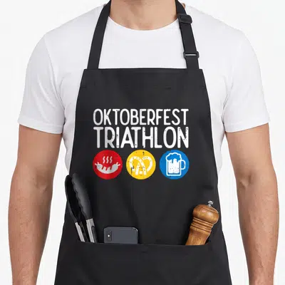 Triathlon oktoberfest apron - Distressed white OKTOBERFEST TRIATHLON text above red sausage, yellow pretzel, and blue — shown on a black cotton canvas apron against a white background