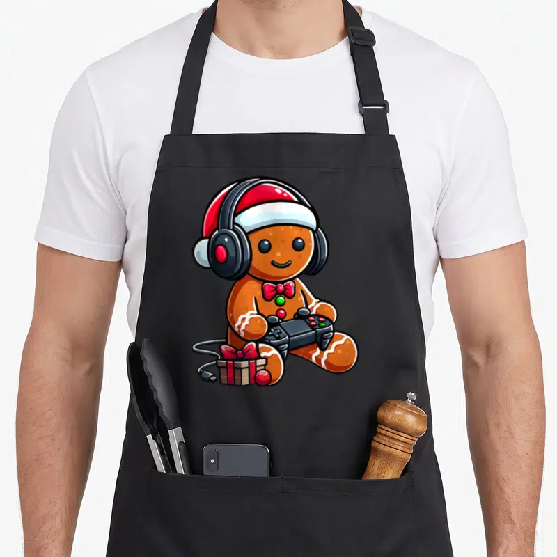 Gamer gingerbread apron - Cartoon gingerbread man with headphones, controller, and gift box in brown, red, white, black — shown on a black cotton canvas apron against a white background
