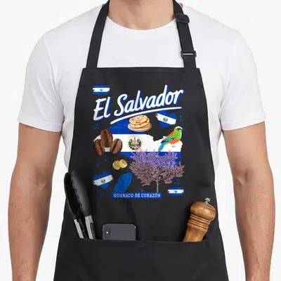 Salvador chef outfit with apron - El Salvador text, pupusas, coffee beans, quetzal bird, and flag elements in blue — shown on a black cotton canvas apron against a white background