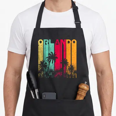 Vacation souvenir florida apron - Retro ORLANDO text with grey palm trees over yellow teal red and orange vertical — shown on a black cotton canvas apron against a white background