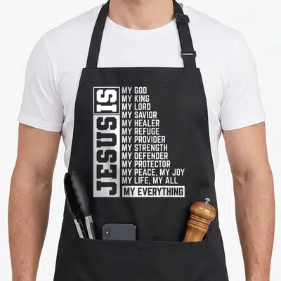 Lord christian aprons - White vertical Jesus Is text beside horizontal list of divine attributes on dark background — shown on a black cotton canvas apron against a white background