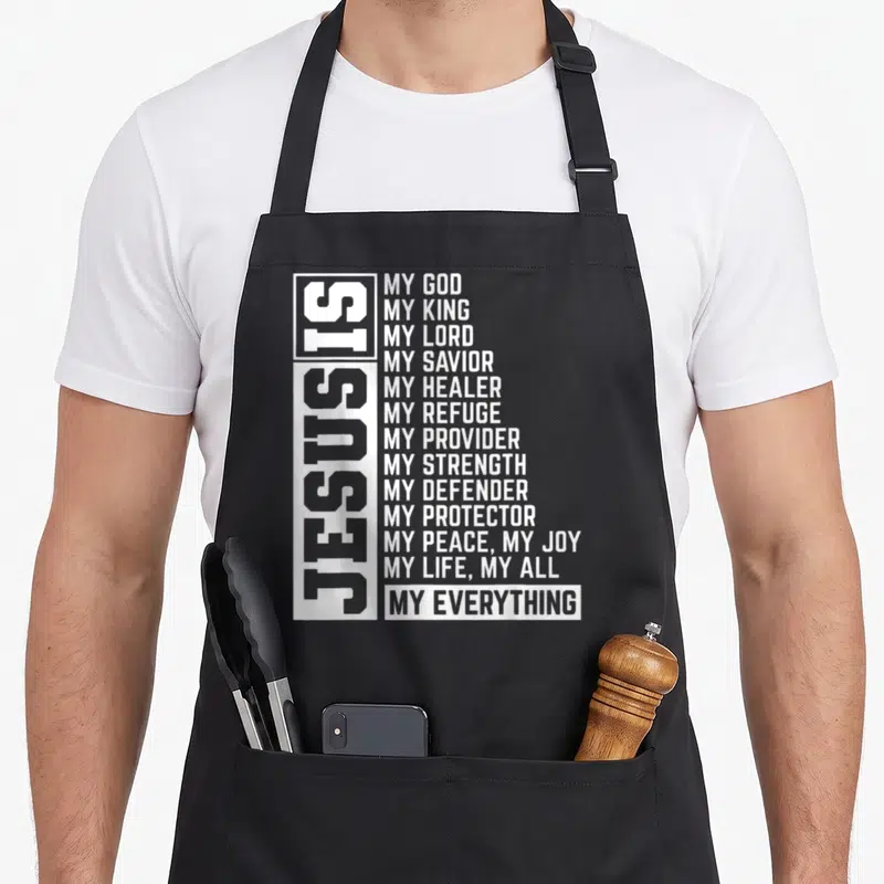 Lord christian aprons - White vertical Jesus Is text beside horizontal list of divine attributes on dark background — shown on a black cotton canvas apron against a white background