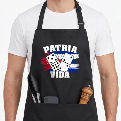 Cubano freedom of movement apron - Patria Y Vida text with crossed dominoes over a distressed flag in white blue and red — shown on a black cotton canvas apron against a white background