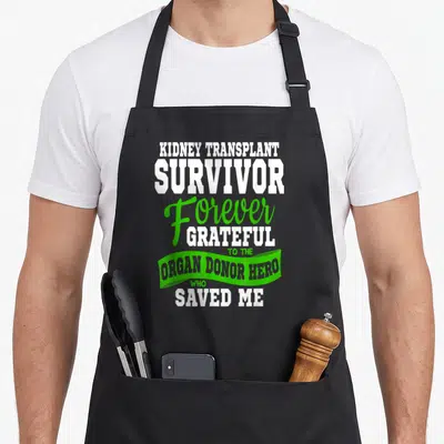 Kidney organ apron - White and green typography reading kidney transplant survivor forever grateful to organ donor hero — shown on a black cotton canvas apron against a white background