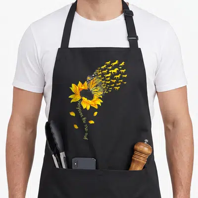 Horse sunflower apron - Sunflower with butterfly turning into flying horses and vertical You are my sunshine text — shown on a black cotton canvas apron against a white background