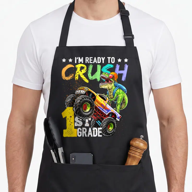1st grade truck apron - T-Rex with backpack standing next to monster truck and colorful text — shown on a black cotton canvas apron against a white background