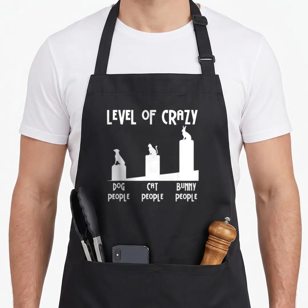 Level bunny apron - White silhouettes of dog cat and bunny on pedestals labeled with text reading level of crazy — shown on a black cotton canvas apron against a white background