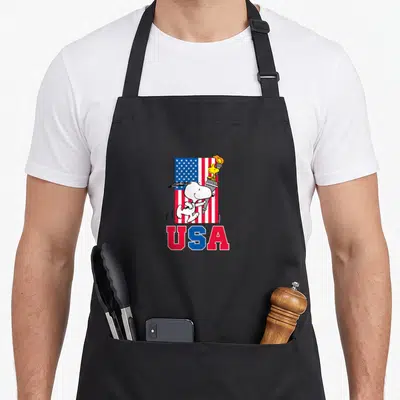 Funny woodstock flag apron - Snoopy running with torch and Woodstock on US flag with USA text below — shown on a black cotton canvas apron against a white background