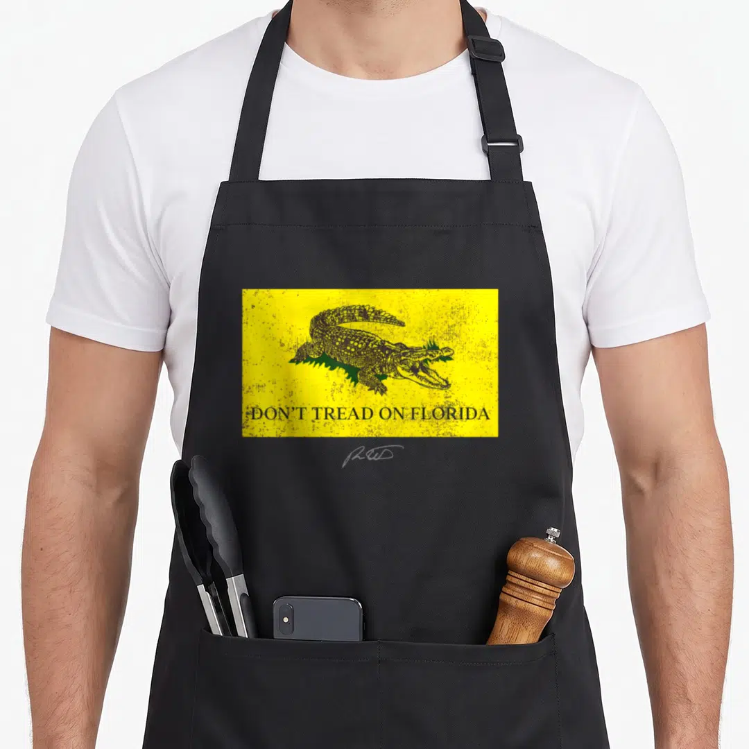 Tread florida apron - Yellow rectangle with black alligator on green grass and text DON'T TREAD ON FLORIDA — shown on a black cotton canvas apron against a white background