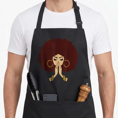 Living magic apron - Minimalist woman with large dark brown afro, tan skin, yellow hoop earrings, and hands in prayer — shown on a black cotton canvas apron against a white background