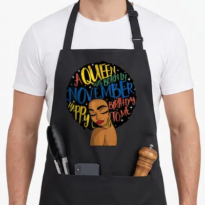 Afro queen birthday apron - Cartoon woman with afro hair and A Queen Was Born In November Happy Birthday To Me text — shown on a black cotton canvas apron against a white background