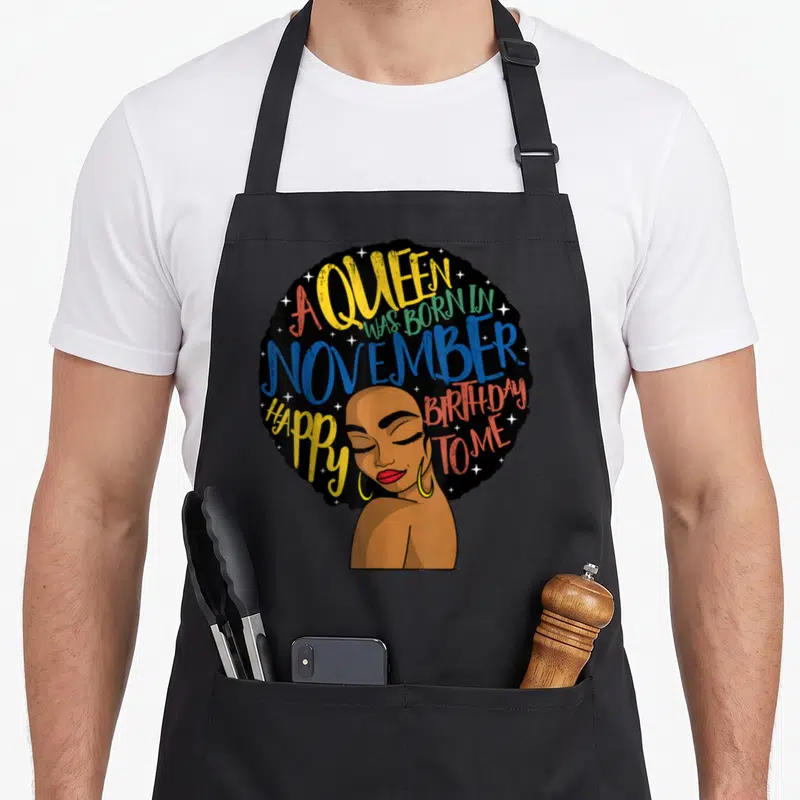 Afro queen birthday apron - Cartoon woman with afro hair and A Queen Was Born In November Happy Birthday To Me text — shown on a black cotton canvas apron against a white background