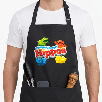 Bottomless veggie apron - Four cartoon hippos in green orange blue and yellow surrounding the Hungry Hungry Hippos logo — shown on a black cotton canvas apron against a white background