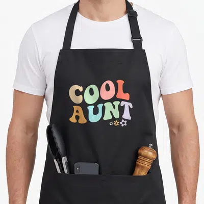Aunt floral apron - Stacked COOL AUNT text in retro colors with three small flowers below the T — shown on a black cotton canvas apron against a white background