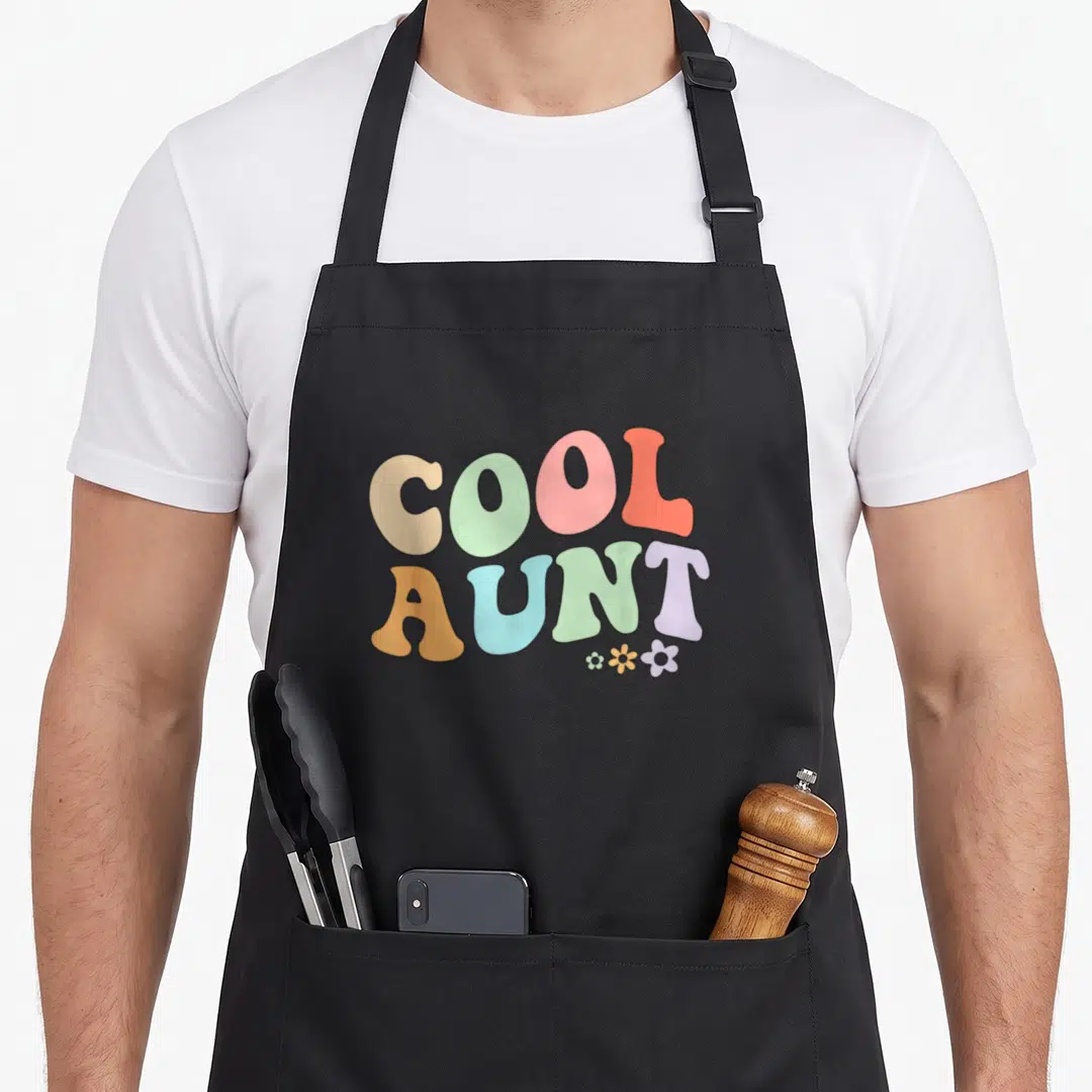 Aunt floral apron - Stacked COOL AUNT text in retro colors with three small flowers below the T — shown on a black cotton canvas apron against a white background