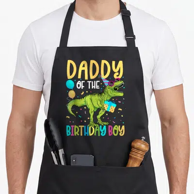 Squad daddy apron - T-Rex with party hat and gift under DADDY OF THE BIRTHDAY BOY text — shown on a black cotton canvas apron against a white background