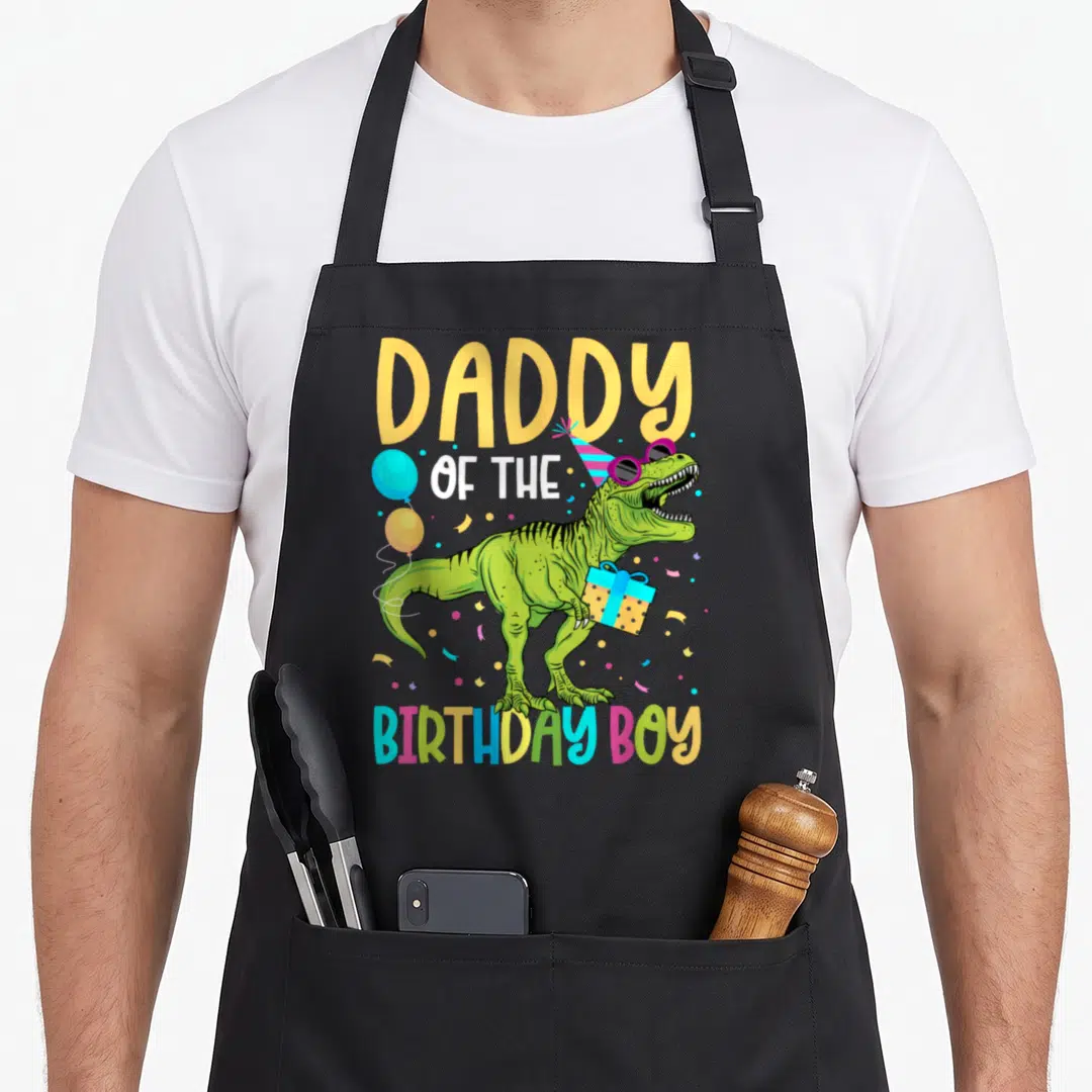 Squad daddy apron - T-Rex with party hat and gift under DADDY OF THE BIRTHDAY BOY text — shown on a black cotton canvas apron against a white background