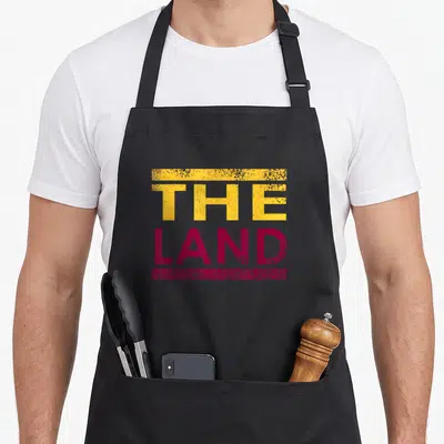 Beach modern apron - THE LAND text in yellow and maroon with distressed horizontal bars — shown on a black cotton canvas apron against a white background