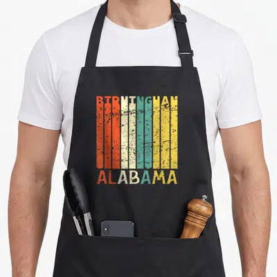Resident alabama apron - Retro BIRMINGHAM ALABAMA text in red cream teal yellow vertical stripes — shown on a black cotton canvas apron against a white background
