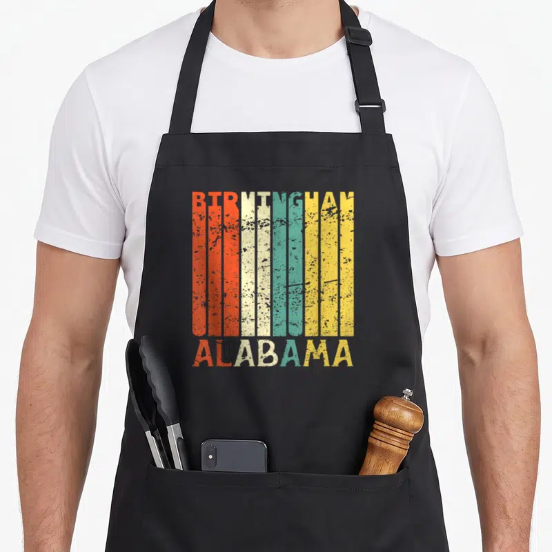 Resident alabama apron - Retro BIRMINGHAM ALABAMA text in red cream teal yellow vertical stripes — shown on a black cotton canvas apron against a white background