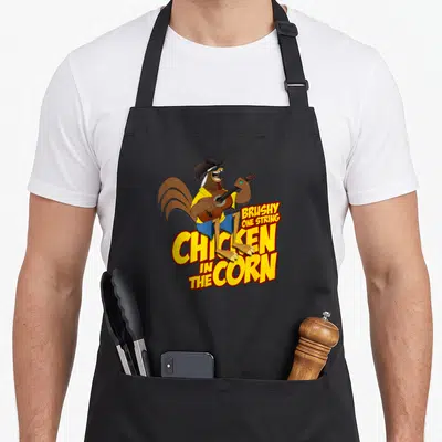 Brushy chicken apron - Cartoon rooster playing guitar above yellow text reading Brushy One String Chicken In The Corn — shown on a black cotton canvas apron against a white background