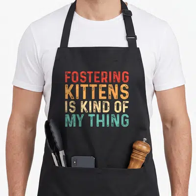 Fostering cat apron - Stacked text reading fostering kittens is kind of my thing in red orange beige and teal — shown on a black cotton canvas apron against a white background