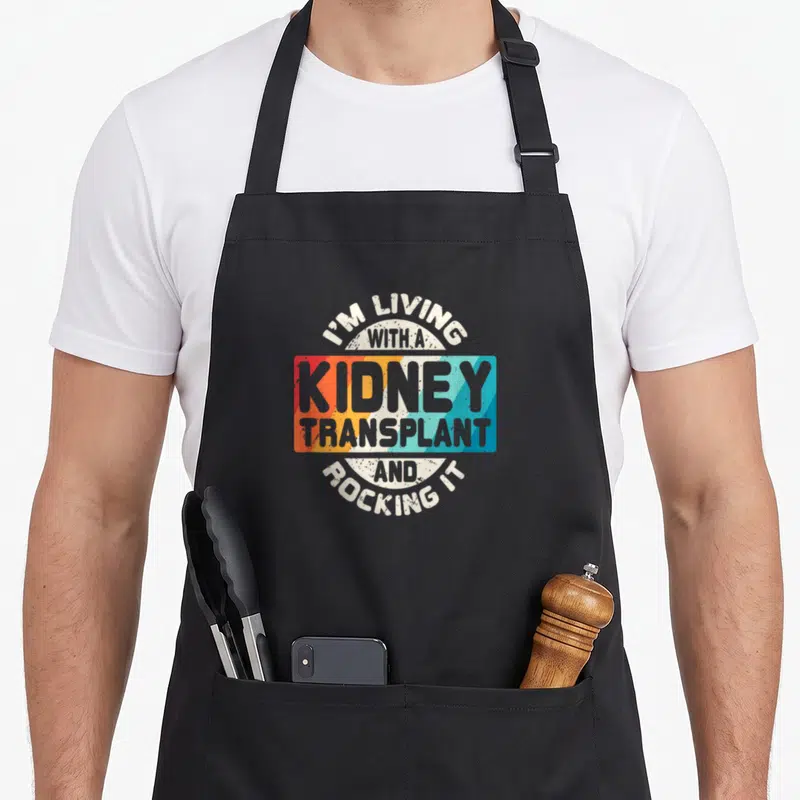 Recipient organ apron - Retro typography reading I'M LIVING WITH A KIDNEY TRANSPLANT AND ROCKING IT over rainbow stripes — shown on a black cotton canvas apron against a white background