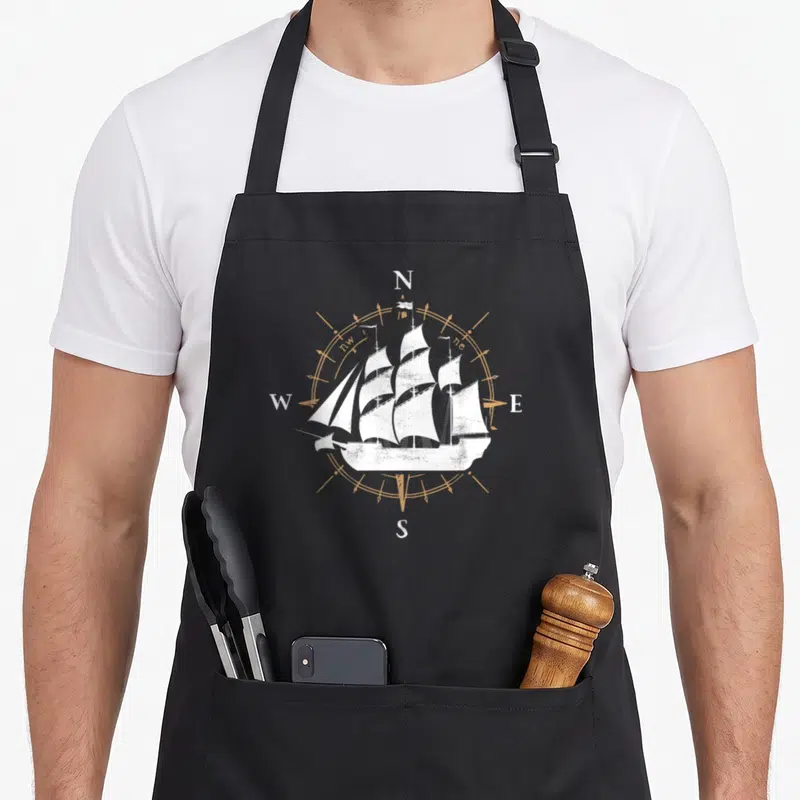 Compass sailfish apron - White sailing ship over tan compass rose with N E S W letters in distressed style — shown on a black cotton canvas apron against a white background