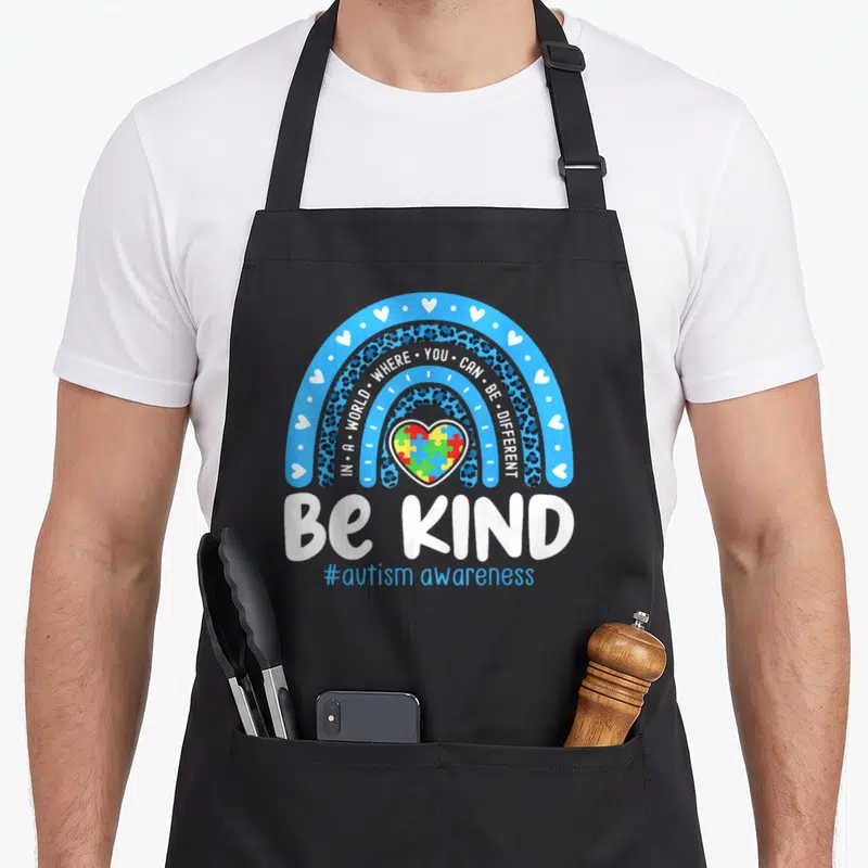 Awareness april apron - Blue rainbow with leopard print arches over a colorful puzzle heart and white Be Kind text — shown on a black cotton canvas apron against a white background