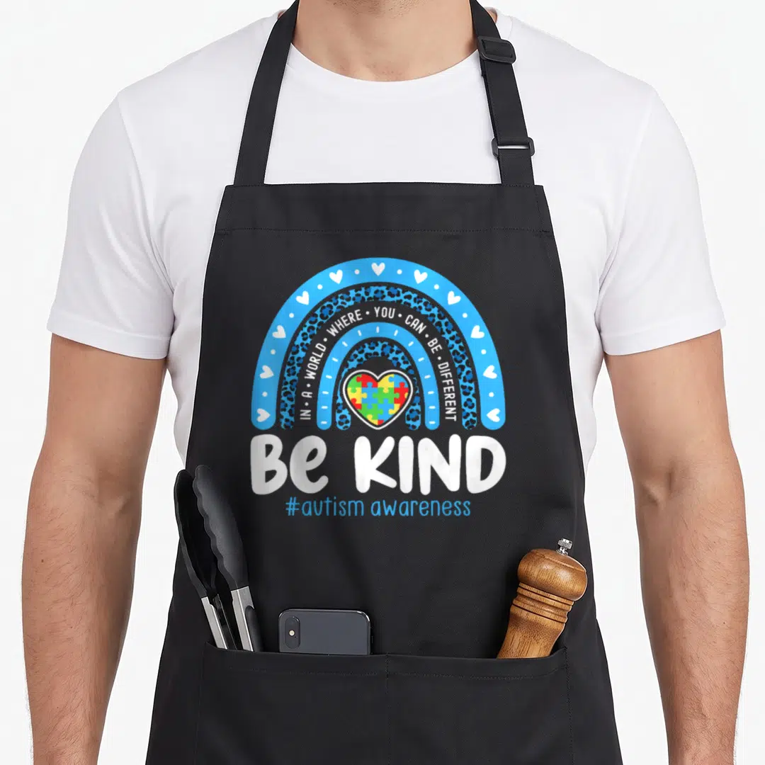 Awareness april apron - Blue rainbow with leopard print arches over a colorful puzzle heart and white Be Kind text — shown on a black cotton canvas apron against a white background