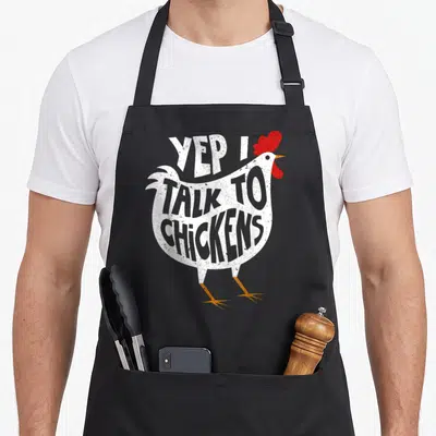 Yep chicken apron - White chicken silhouette containing YEP I TALK TO CHICKENS text with red comb and orange legs — shown on a black cotton canvas apron against a white background