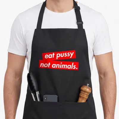 Animal vegan apron - White text eat pussy not animals on stacked red rectangular bars — shown on a black cotton canvas apron against a white background