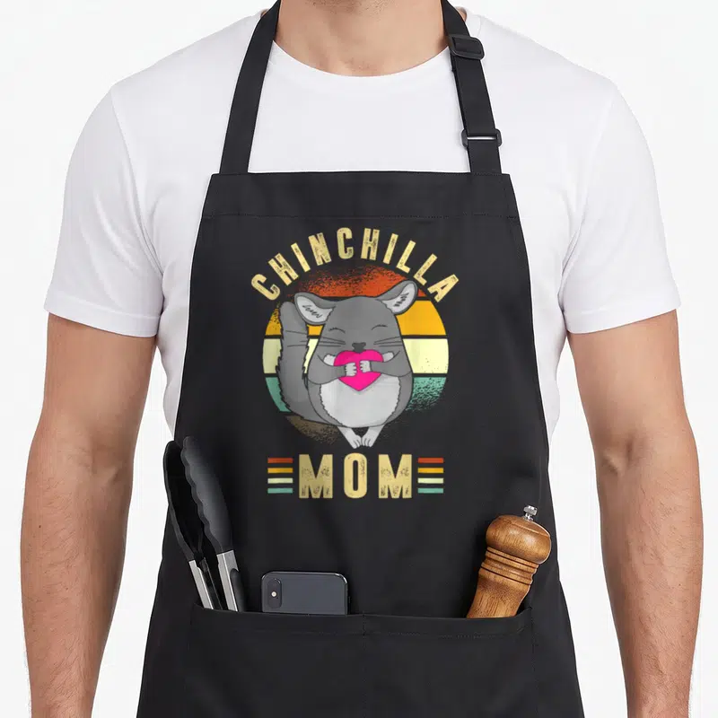 Vintage chinchilla mom apron - Retro grey chinchilla holding pink heart inside orange and teal sunset circle — shown on a black cotton canvas apron against a white background